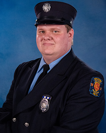 A male firefighter in uniform poses against a blue background. He wears a dark jacket with a badge, a black tie, and a matching cap displaying a silver emblem. A fire department patch is visible on his upper left sleeve.