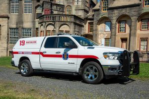 A white fire rescue truck marked "Battalion 19" is parked on grass in front of a large, historic stone building with many windows. The truck has red stripes, an emblem, and chrome wheels.