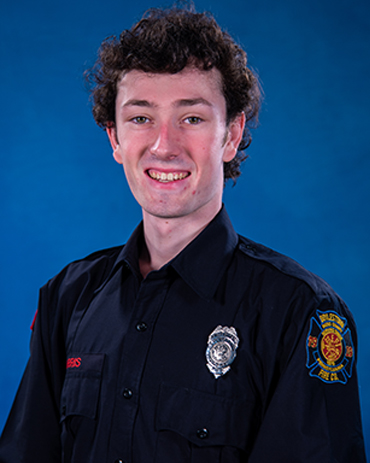 A young man with curly brown hair wearing a dark firefighter uniform, a silver badge, and a patch on the sleeve, smiling in front of a solid blue background.
