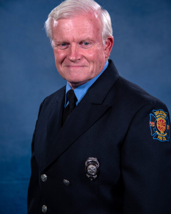 An older man with short white hair wears a dark blue firefighter uniform, featuring a badge on his chest and a fire department patch on his sleeve, posing in front of a blue backdrop.