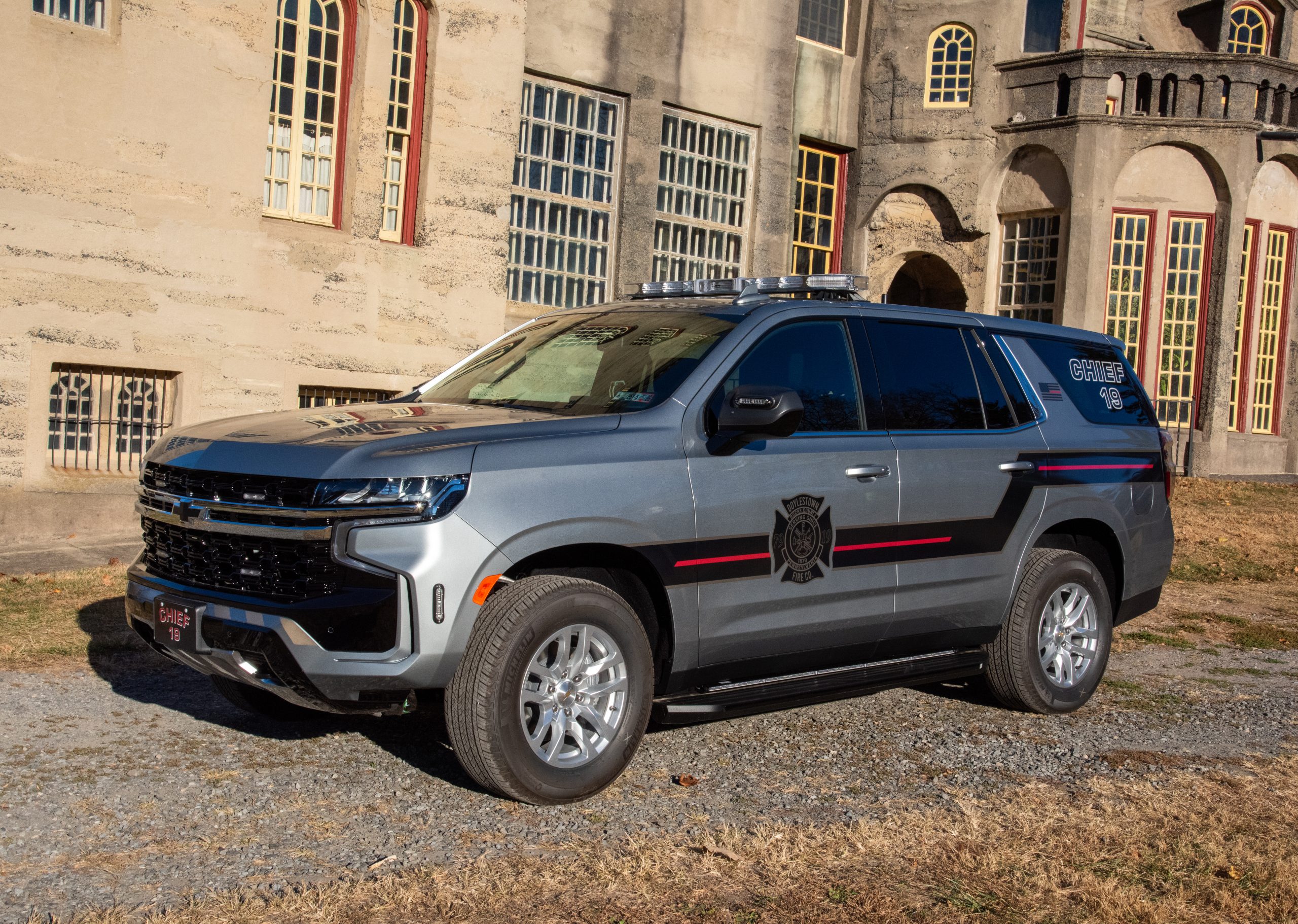 A silver Chevrolet police SUV with black and red stripes is parked on gravel in front of an old, beige stone building with tall windows. The words "RANGER" and a badge emblem are visible on the vehicle's side.