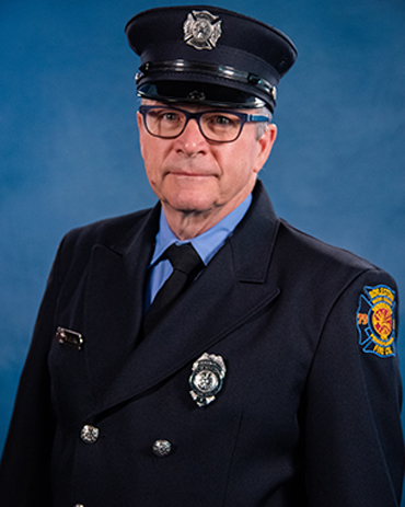 A middle-aged man wearing glasses and a formal dark blue firefighter uniform with badges and a hat, standing in front of a blue background.