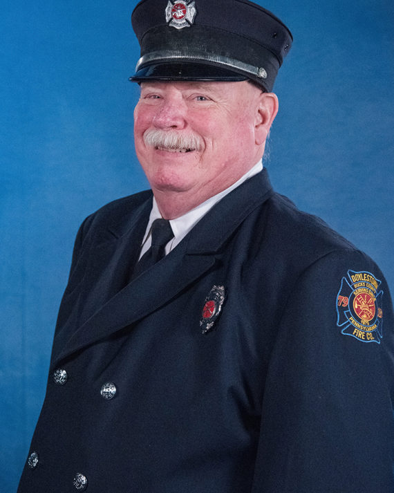A smiling firefighter with a white mustache wearing a dark blue uniform and cap stands in front of a blue background. A fire department badge and an embroidered patch are visible on his uniform.