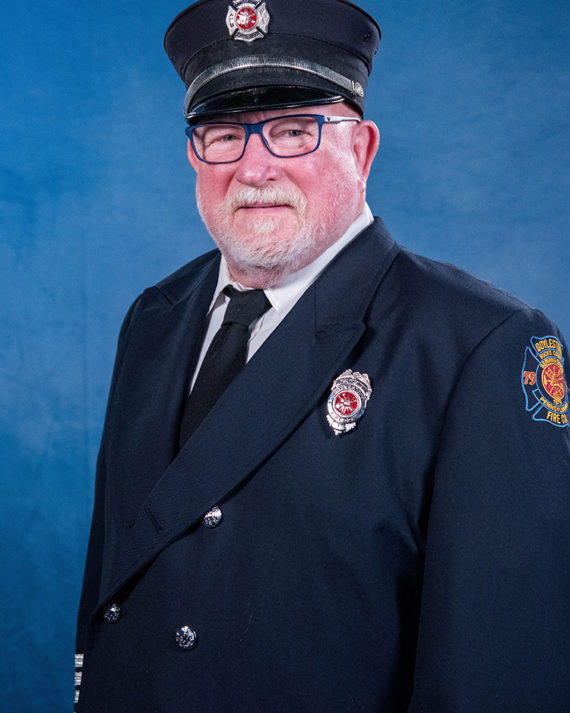 An older man with a white beard and glasses wears a dark blue firefighter dress uniform, a matching hat with an emblem, and a tie, standing in front of a blue background and smiling slightly.