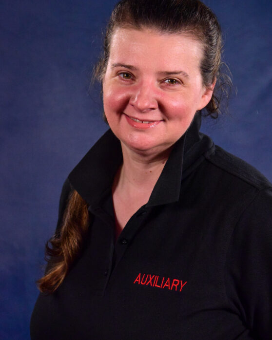 A woman with long brown hair in a ponytail smiles at the camera, wearing a black collared shirt with "AUXILIARY" written in red letters, standing against a blue background.