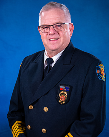 A man with short gray hair and glasses, wearing a dark formal firefighter uniform with medals, buttons, and a department patch, poses in front of a blue background.