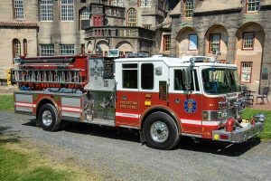 A red and white fire truck from Doylestown Fire Co. No. 1 is parked in front of a large, historic building with arched windows and stone walls on a sunny day.
