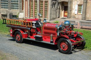 A vintage red fire truck with wooden ladders and chrome details is parked on a gravel path in front of an old building with tall windows and stone walls.