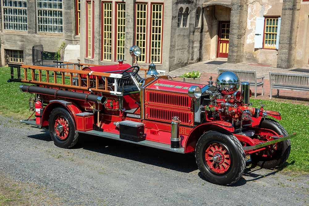 A vintage red fire truck with wooden ladders and chrome details is parked on a gravel path in front of an old building with tall windows and stone walls.