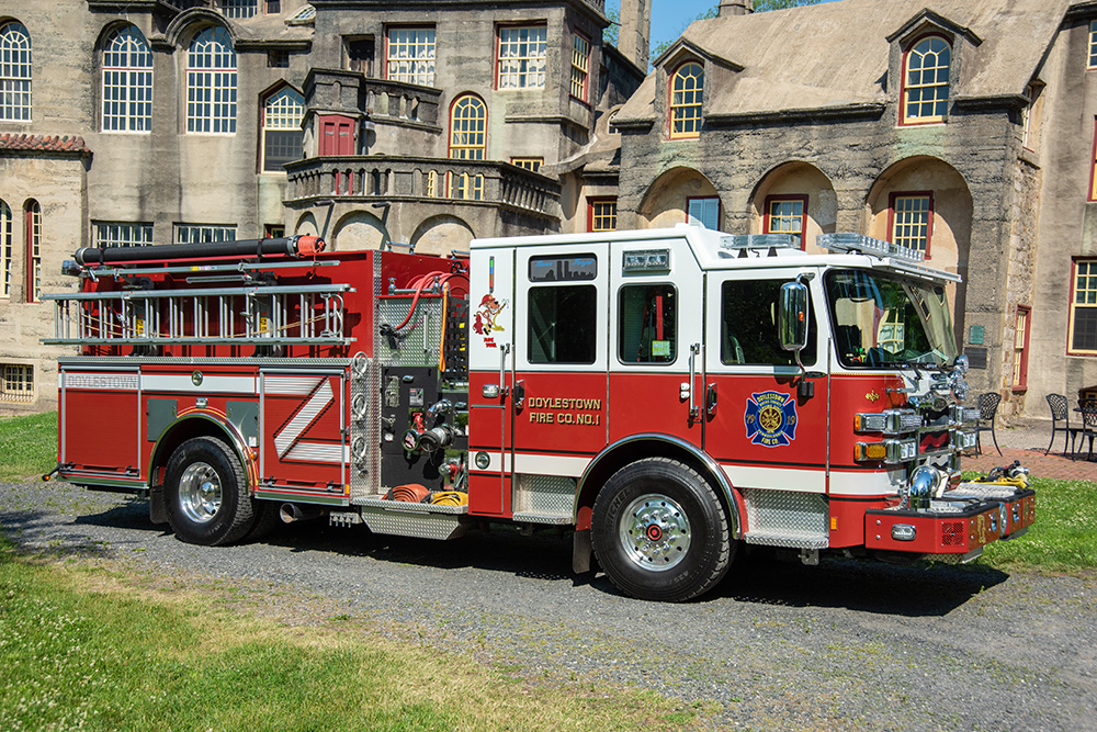 A red and white fire truck from "Doylestown Fire Co. No. 1" is parked on a gravel path in front of a historic, castle-like building with arched windows and textured stone walls.