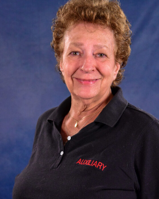 Smiling older woman with short curly hair wears a black polo shirt with "AUXILIARY" embroidered in red, standing in front of a blue backdrop.
