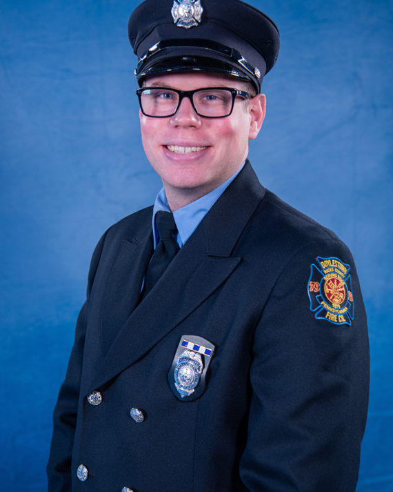 A smiling firefighter in formal uniform with a hat, glasses, and a badge, stands in front of a blue background. The patch on their sleeve reads "Coplay Fire Co." with an emblem.