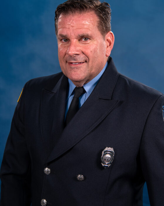 A man in a dark formal uniform with a badge on his chest stands against a blue background, smiling at the camera.
