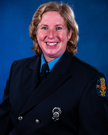 A smiling woman with short, curly blonde hair wears a dark blue formal firefighter uniform with a badge and an embroidered patch on the sleeve, posed in front of a blue background.