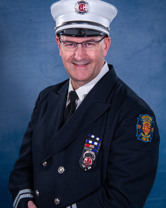 A smiling firefighter in formal dress uniform and hat poses against a blue backdrop. He wears glasses, a badge, medals, and a patch on his sleeve that reads "FIRE DEPT.