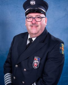 A man wearing a dark formal firefighter uniform and hat, with badges and insignia, poses in front of a blue background. He has glasses, a mustache, and is smiling at the camera.