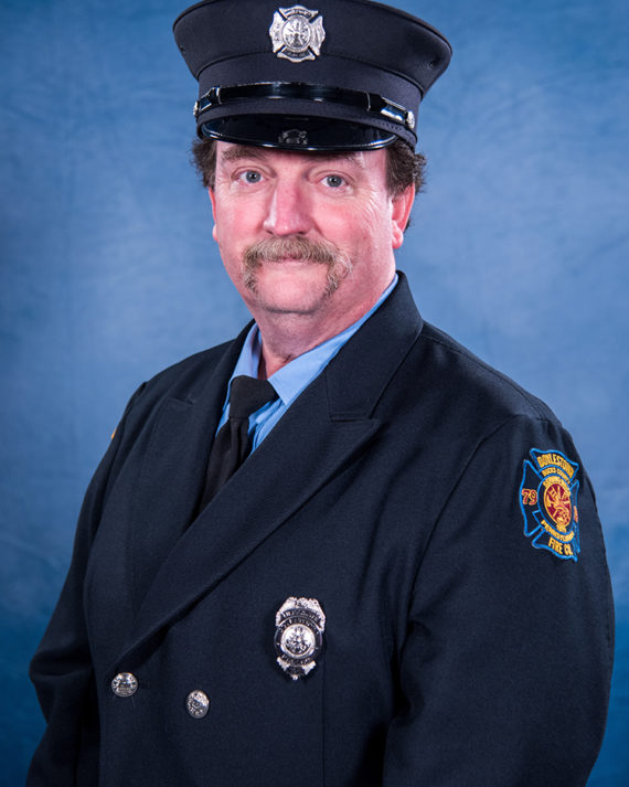 A firefighter with a mustache wearing a formal navy blue uniform and cap poses in front of a blue background. A fire department badge and patch are visible on his jacket.