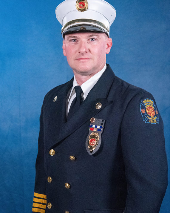A firefighter in a formal dark blue uniform with gold buttons and badges stands in front of a blue backdrop. He wears a white peaked cap and a medal pinned to his chest.