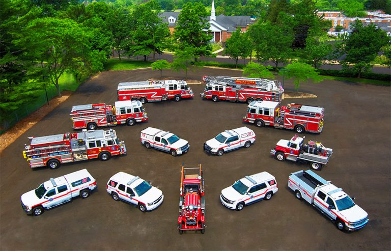An aerial view of various fire department vehicles, including fire trucks, SUVs, pickup trucks, and a vintage fire engine, arranged in a semicircle on a paved area surrounded by trees and buildings.