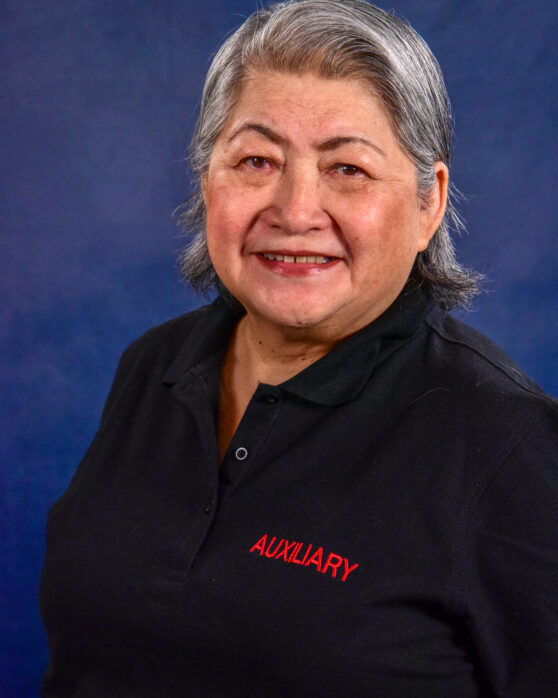 A smiling older woman with gray hair wearing a black polo shirt that says "AUXILIARY" in red letters stands in front of a blue background.