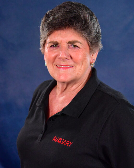 An older woman with short gray hair smiles at the camera. She is wearing a black collared shirt with the word "AUXILIARY" embroidered in red on the chest. The background is a plain blue backdrop.