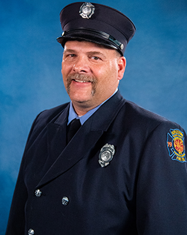 A man in a dark blue firefighter uniform and hat, wearing a badge and smiling, stands in front of a blue background.