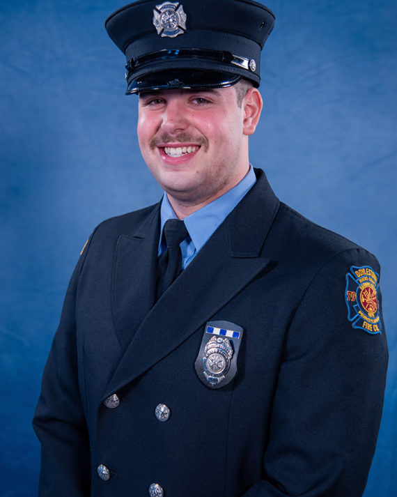 A smiling firefighter in a formal dark blue uniform and hat stands against a blue background. A badge and patch are visible on his uniform, and he has a moustache.