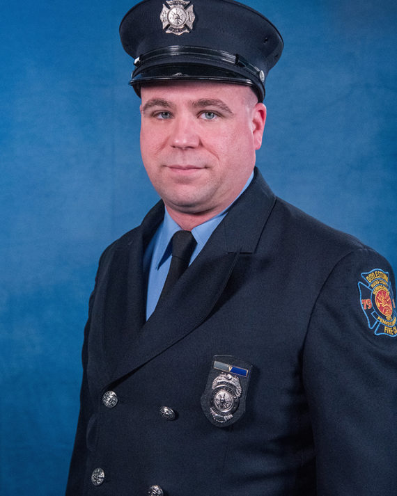 A firefighter in formal dress uniform with a badge and hat poses in front of a blue background.