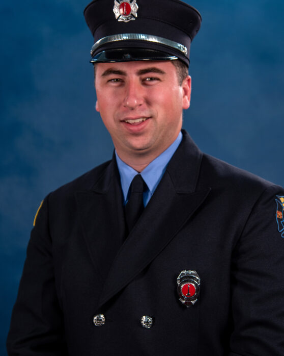 A man dressed in a dark blue firefighter uniform and hat poses in front of a blue backdrop, smiling slightly. A badge and department patch are visible on his jacket and sleeve.