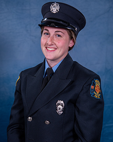 A smiling firefighter in full dress uniform, featuring a dark jacket with badges, a tie, and a hat with an emblem, stands in front of a blue background.