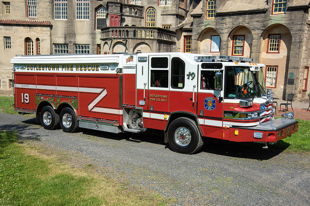 A red and white Doylestown Fire Rescue truck is parked on a grassy and gravel area in front of a large, historic stone building with arched windows and decorative architectural details.