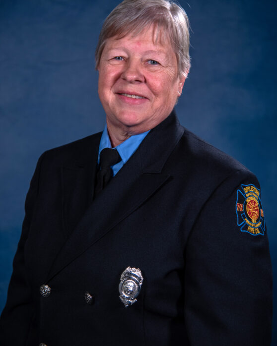 An older woman with short gray hair smiles while wearing a dark firefighter uniform with a badge, patch, and tie, posing in front of a blue background.