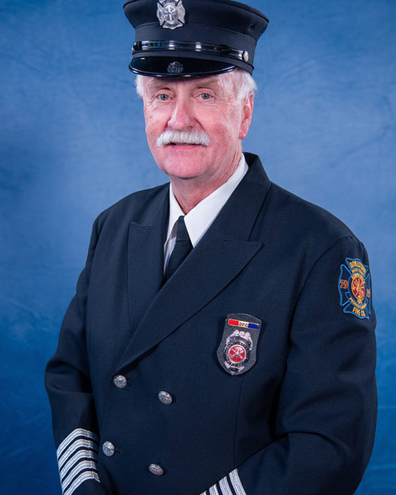 An older man with white hair and a mustache wears a dark firefighter’s dress uniform and cap, standing in front of a blue background and facing the camera with a slight smile.