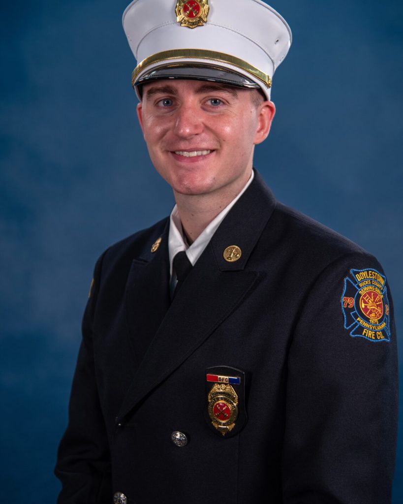 A young firefighter in formal dress uniform and white hat poses against a blue background, smiling at the camera. The uniform features badges and a shoulder patch that reads "Doylestown Fire Co.