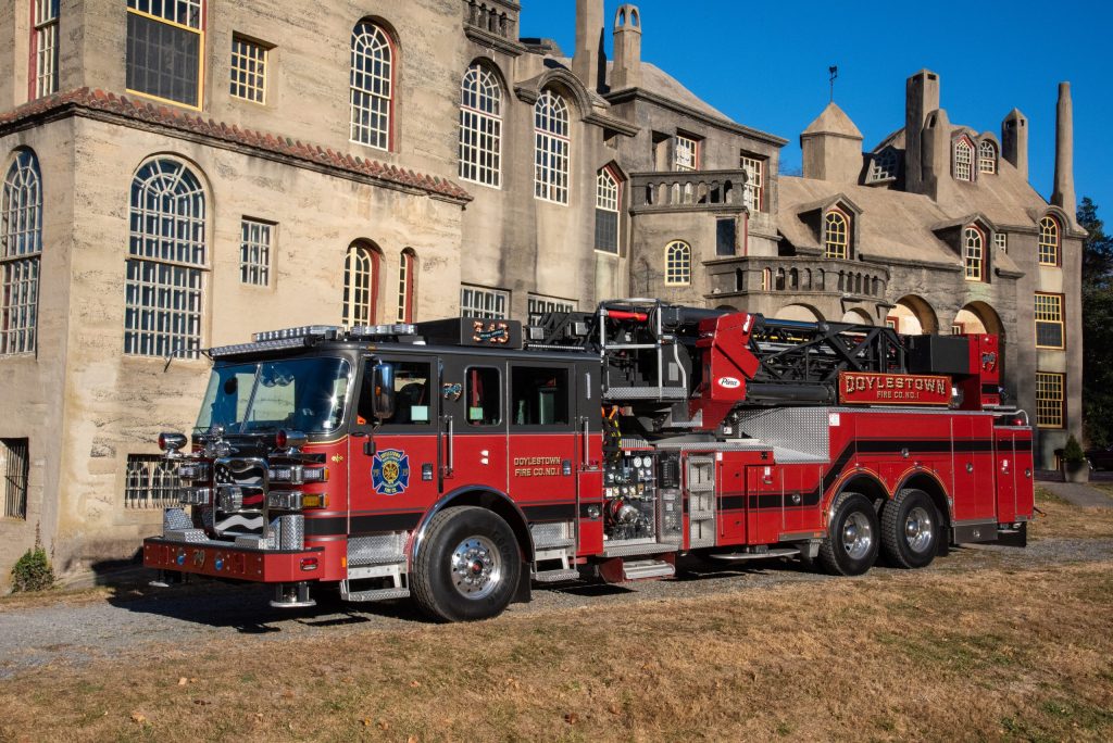 A red and black fire truck labeled “Doylestown” is parked on dry grass in front of a large, historic stone building with tall windows and castle-like architecture.