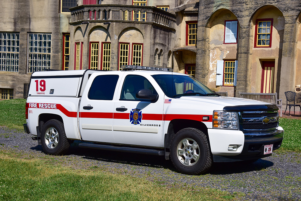 A white fire rescue pickup truck with red stripes and "19 TRAFFIC" markings is parked outside a historic building with yellow and red window frames.