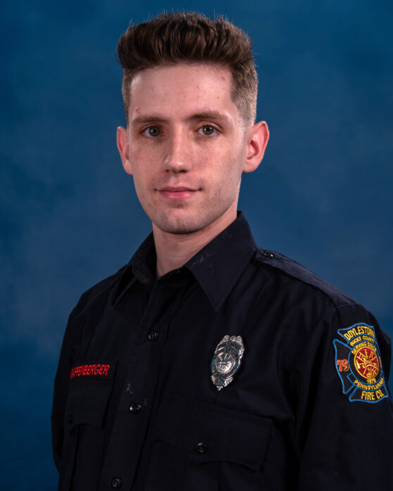 A young man in a dark firefighter uniform with a badge and embroidered name stands against a plain blue background, looking at the camera with a neutral expression.