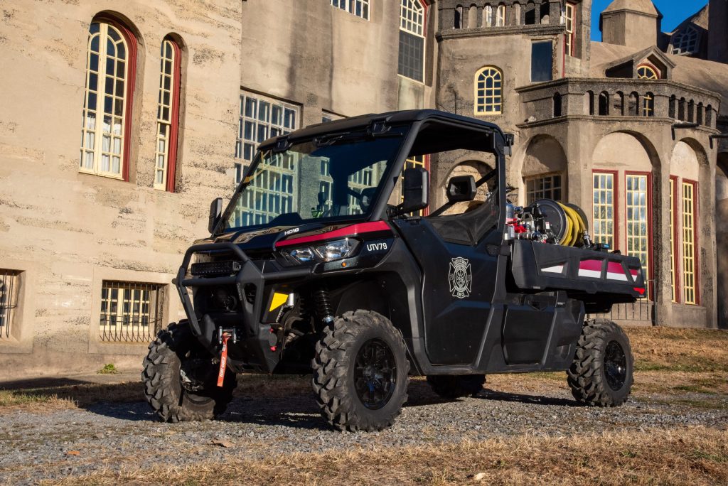 A black utility terrain vehicle (UTV) with emergency equipment mounted on the back is parked on gravel outside a large, historic building with tall windows and beige walls.