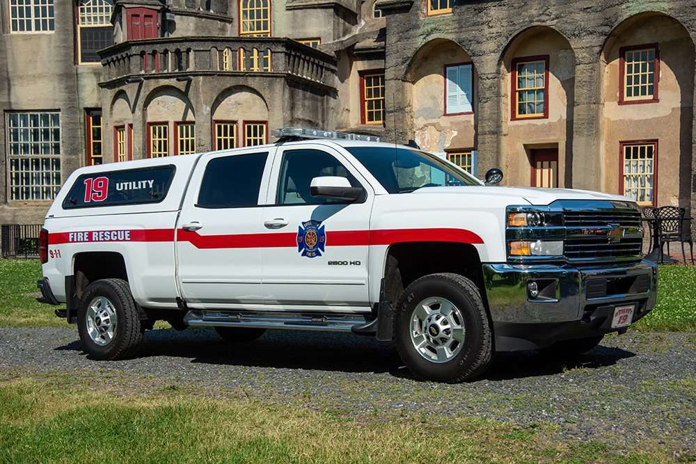 A white fire rescue utility truck with red stripes and the number 19 is parked on grass in front of an old stone building with arched windows.