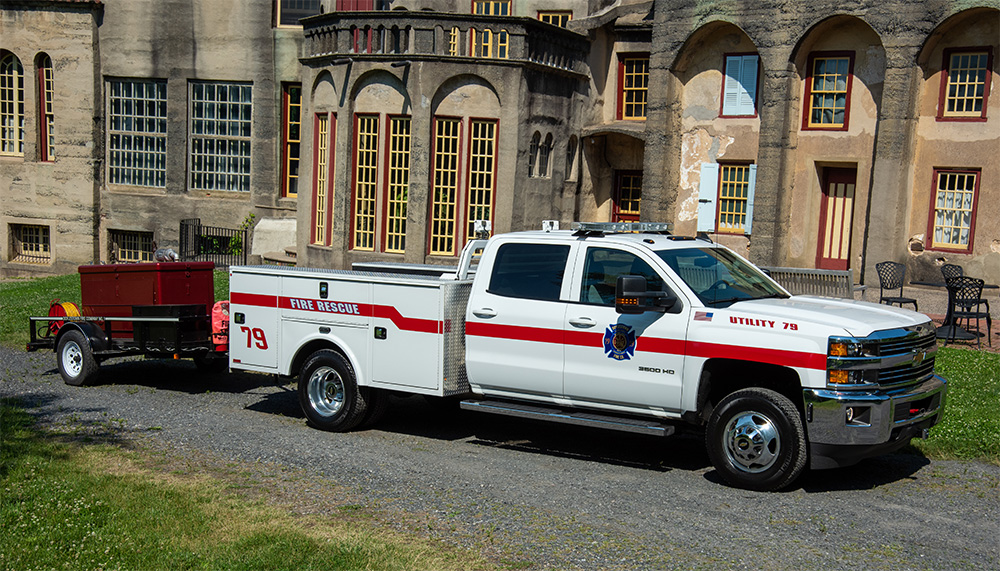 A white fire rescue utility truck with red stripes and the number 79 is parked outside a historic building, towing a small red trailer.
