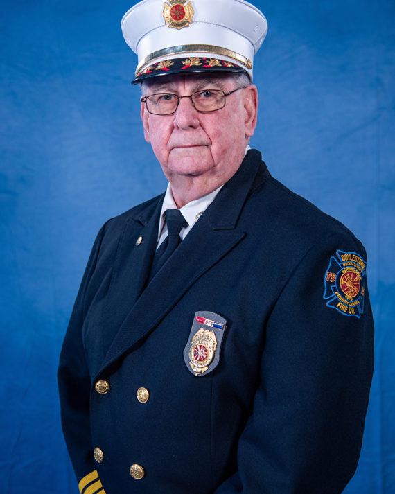An older man in a formal firefighter dress uniform and hat stands in front of a blue background, looking at the camera with a neutral expression. Badges and an embroidered patch are visible on his jacket.