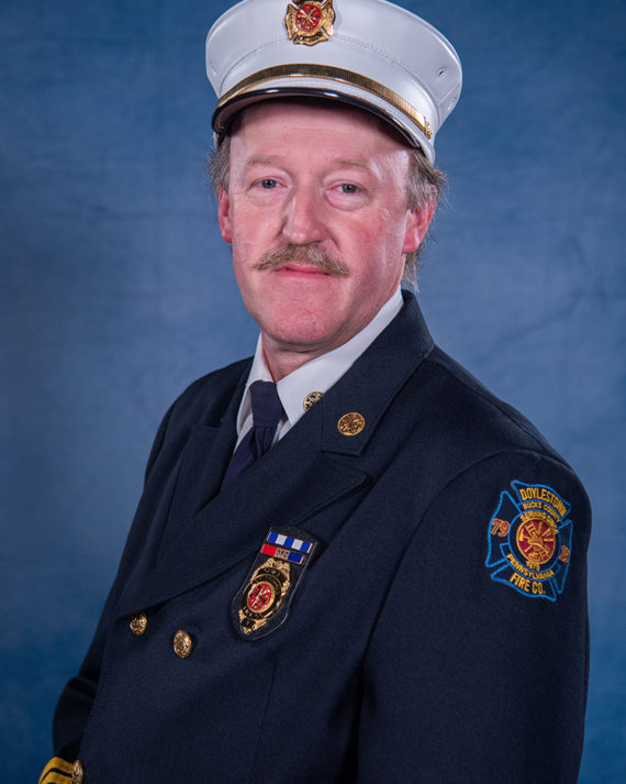 A man in a formal firefighter uniform and white hat poses against a blue background. He has a mustache and wears a dark jacket with badges and a patch reading “Doylestown Fire Co.” on the sleeve.