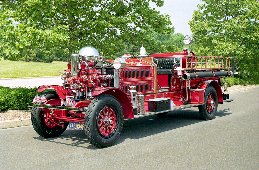 A shiny, vintage red fire truck with chrome details and ladders is parked on a paved road, surrounded by green trees and bushes on a sunny day.