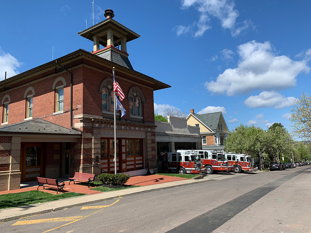 A historic brick fire station with an American flag, fire trucks parked outside, and a tree-lined street under a blue sky with scattered clouds.