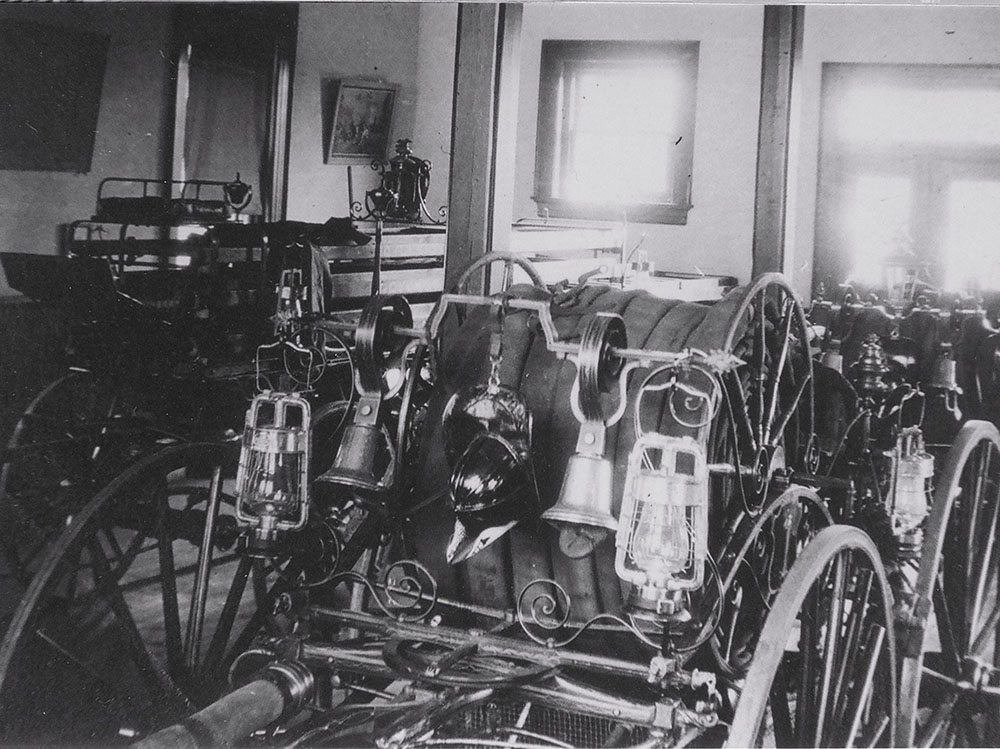 Black and white photo showing antique fire wagons with large wooden wheels, lanterns, helmets, and bells, arranged inside a building with light streaming through windows.