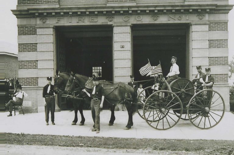 Historic black-and-white photo of firefighters in uniform with a horse-drawn fire wagon outside a brick fire station. An American flag is held by a firefighter seated on the wagon.