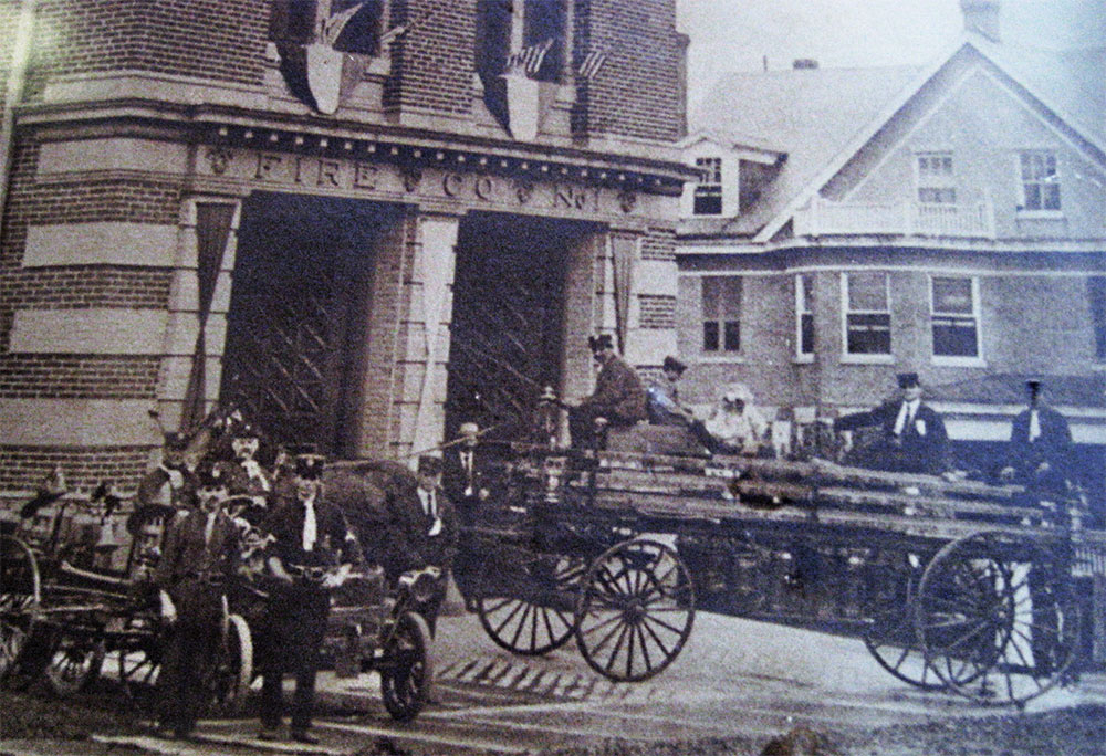 Vintage black-and-white photo of firefighters with horse-drawn fire wagons outside a brick firehouse. Several men in uniforms pose with equipment; the building behind has "FIRE CO. NO. 1" above the entrance.