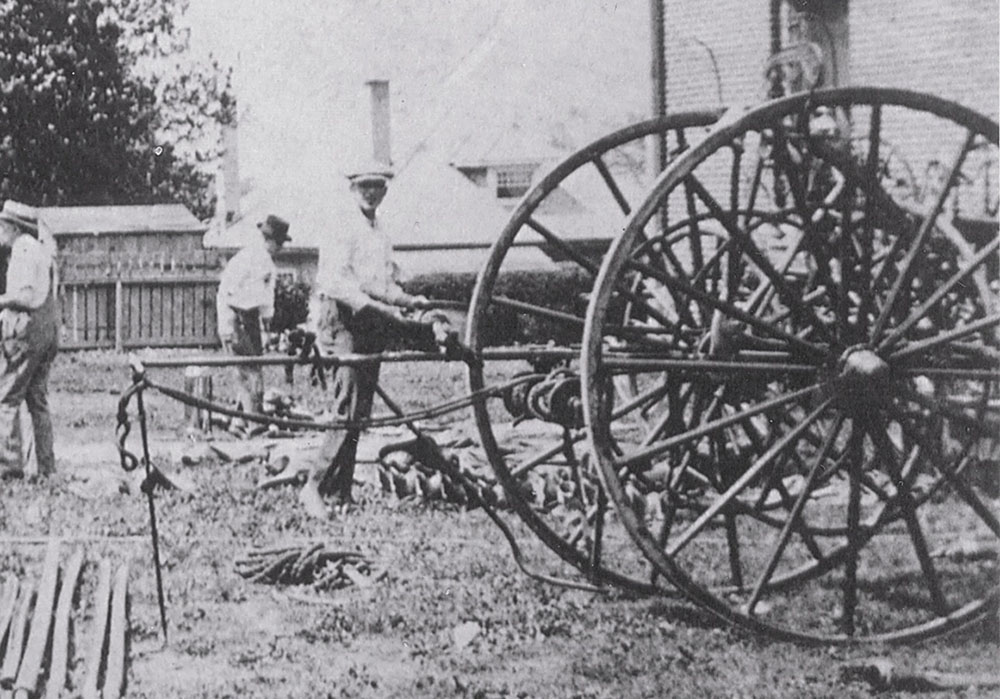 Three men in hats stand in a yard next to a large, old-fashioned wheeled fire engine or hose reel, with tools and equipment lying on the ground nearby. The scene appears historical, possibly from the early 1900s.