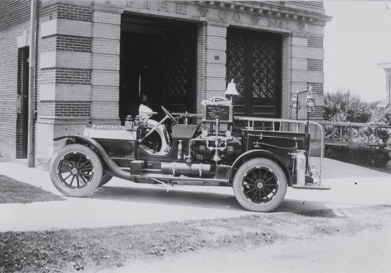 A vintage fire truck is parked outside a brick fire station with large, dark double doors. The truck features ladders, hoses, and firefighting equipment on its sides.