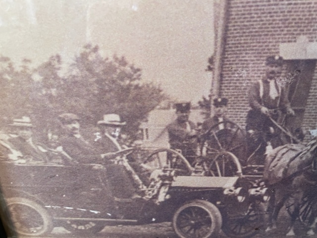 A sepia-toned vintage photograph shows several men in an old-fashioned car and others on a horse-drawn carriage in front of a brick building, with trees visible in the background.